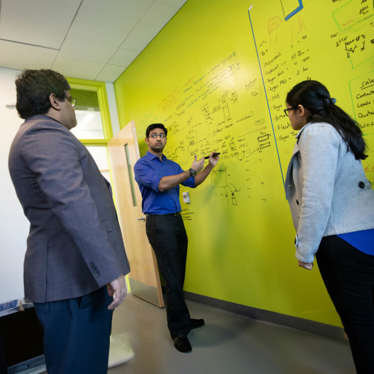 Three people discuss and review an elaborate system of notes on a wall-sized whiteboard.