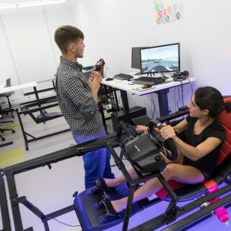 One student observes a monitor displaying first-person view from an automobile as another navigates inside an artificial cockpit.