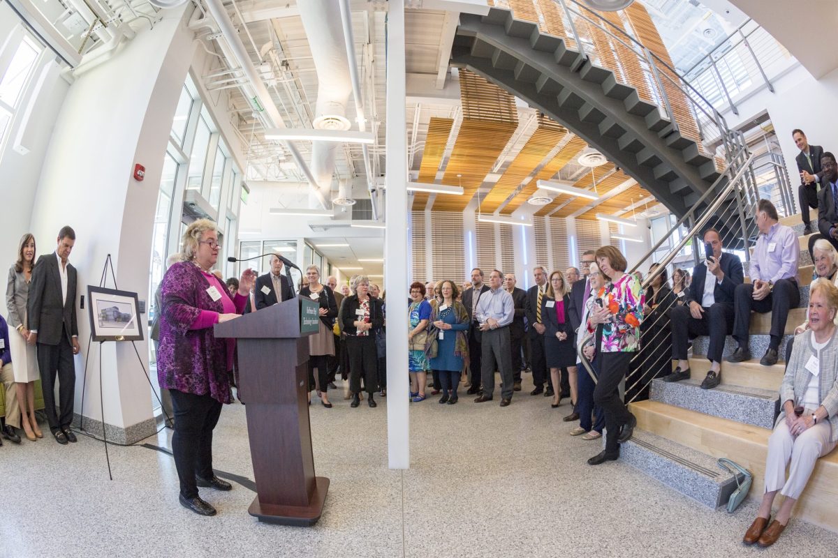 A speaker at a podium presents to a large group of people seated throughout a lobby area in the Koffman building.