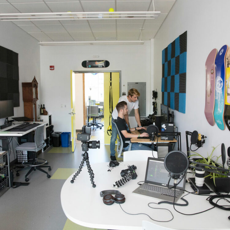 Two students look at a laptop screen in a room full of AV equipment.